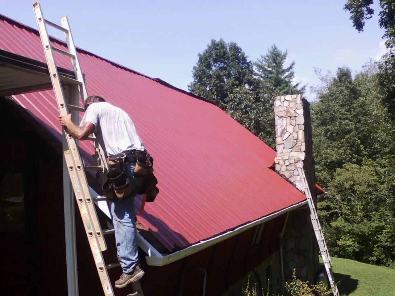 Roofer on a ladder beside a red metal roof and stone chimney — Soffit & Fascia Repair by Roofing Brothers in Mallory
